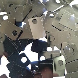 Könnte beinhalten: Eine Ansammlung kleiner, schwarzer, rechteckiger Metallanhänger mit einem kreisförmigen Loch oben. Die Anhänger sind auf einer weißen Oberfläche verstreut und erzeugen einen strukturierten, geschichteten Effekt. Die Anhänger haben eine glänzende, reflektierende Oberfläche.