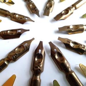 May include: A collection of vintage calligraphy pen nibs in various shapes and sizes, arranged in a circular pattern. The nibs are made of brass and have intricate designs. Some have the brand name "LEON" visible.