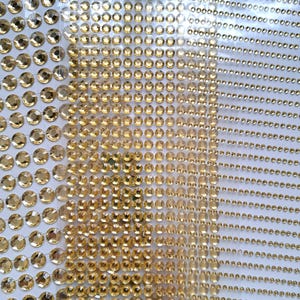 May include: Sheets of golden, faceted rhinestones in various sizes. The gems are arranged in rows and columns, and are adhered to a clear, adhesive backing. The rhinestones have a reflective surface, and are suitable for crafts and embellishments.