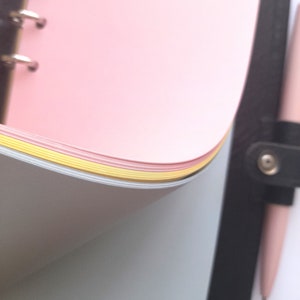 May include: A pink, blue and yellow notebook with a black leather cover and a silver ring binder. The notebook is open to reveal the different coloured pages.
