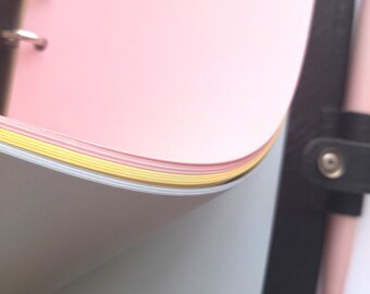 May include: A pink, blue, and yellow notebook with a black leather cover and a silver ring binder. The notebook is open to reveal the different colored pages.