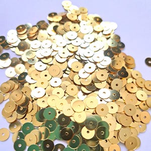 May include: A collection of gold-coloured sequins, each with a central hole. The sequins are scattered, creating a textured effect. These small, circular embellishments are suitable for various craft projects and add a touch of sparkle.