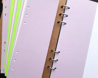 May include: Open black binder with a stack of pastel-colored, hole-punched paper inserts. The inserts include shades of pink, green, and white. The binder is open, revealing the metal rings and a blank white page with a dotted pattern.