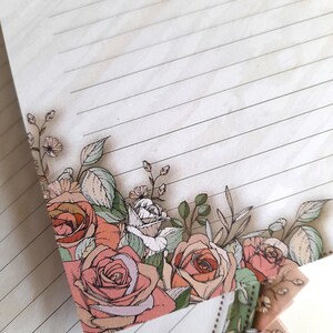 May include: A stack of lined paper with a floral design on the bottom edge. The design features pink and white roses, green leaves, and small white flowers.