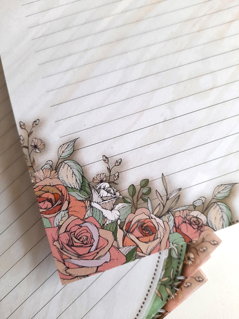 May include: A stack of lined paper with a floral design on the bottom edge. The design features pink and white roses, green leaves, and small white flowers.