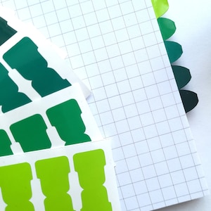 May include: A sheet of grid paper with green and lime green sticker tabs. The tabs are in different shades of green and are arranged in a fan shape.
