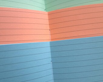 May include: An open notebook with pages in shades of blue, orange, and green. Each page has horizontal black lines for writing. The notebook is bound with white thread.