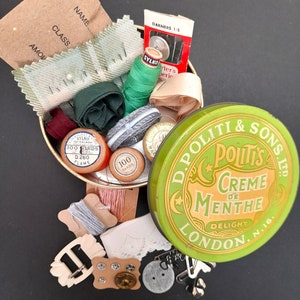 Könnte beinhalten: Eine grüne Metalldose mit einem weißen Halbmond und dem Text "D. Politi & Sons Ltd. Poult's Creme de Menthe Delight London N.16." Die Dose ist gefüllt mit Nähzubehör, darunter Garnrollen, Knöpfe und Nadeln.