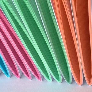 May include: A stack of colorful notebooks with white edges. The notebooks are arranged in a fan shape, with the covers facing outward. The colors of the notebooks are blue, pink, green, and orange.