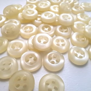May include: A collection of off-white plastic buttons with four holes each. The buttons are arranged in a random pattern on a white surface.