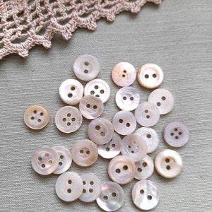 May include: A collection of white mother of pearl buttons with two or four holes. The buttons are arranged in a random pattern on a light grey fabric background.
