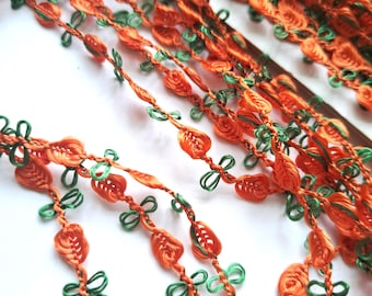 May include: A decorative trim featuring a repeating pattern of orange, leaf-shaped elements interspersed with small, looped green thread accents. The trim is constructed from a series of interconnected, woven orange and green fiber strands.