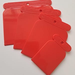 May include: A set of red plastic scrapers in different sizes. Each scraper features a rectangular blade with rounded edges and a textured handle with a hanging hole. The scrapers are arranged overlapping each other.