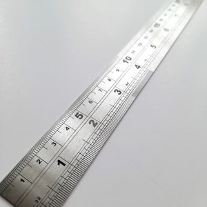 May include: A silver metal ruler with inch and centimetre markings. The ruler is 12 inches long and has markings for every 1/32 of an inch. The ruler is on a white surface.