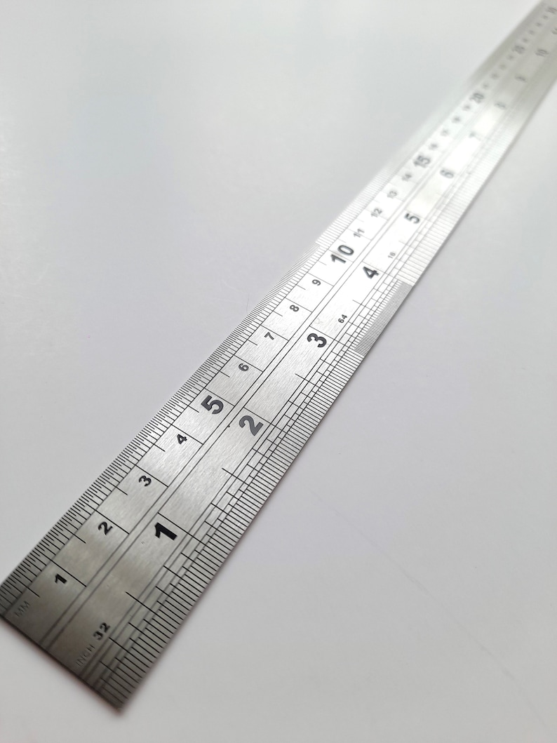 May include: A silver metal ruler with inch and centimetre markings. The ruler is 12 inches long and has markings for every 1/32 of an inch. The ruler is on a white surface.