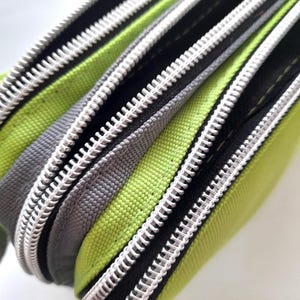 May include: Close-up of three zippered pouches in shades of green, grey, and black. The zippers are silver and the fabric appears to be a durable canvas material. The pouches are partially open, revealing the interior.