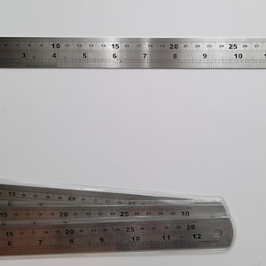 May include: Four silver metal rulers stacked on top of each other. The rulers are marked in inches and have black numbers. The top ruler is 12 inches long, and the bottom ruler is 30 inches long.