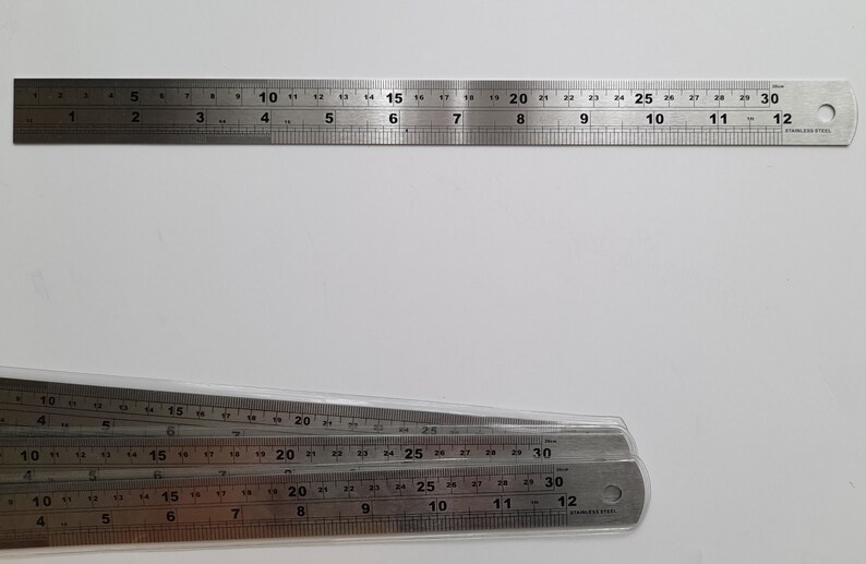 May include: Four silver metal rulers stacked on top of each other. The rulers are marked in inches and have black numbers. The top ruler is 12 inches long, and the bottom ruler is 30 inches long.