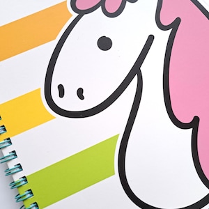 Könnte beinhalten: Ein Spiralnotizbuch mit einem Einhorn-Design. Das Einhorn hat eine rosa Mähne und eine gelbe Krone, die schwarz umrandet sind. Das Cover des Notizbuchs hat farbenfrohe horizontale Streifen in Gelb, Orange, Grün und Türkis. Die Spiralbindung ist türkis.