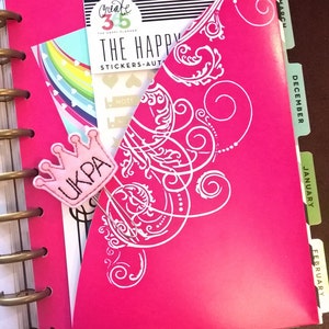 May include: A pink planner with a white decorative design and a pink crown clip with the word "UKPA" on it. The planner has tabs for each month of the year, including January, February, March, December.