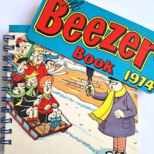 May include: A spiral-bound notebook featuring a colourful comic illustration of children on a sled and a person in a purple coat. The cover includes a blue rectangular overlay with the red and yellow text The Beezer Book 1974 printed on it.