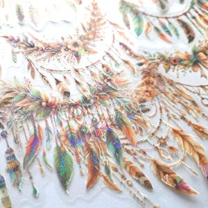 May include: Close-up of decorative stickers featuring dreamcatcher designs. The stickers showcase intricate details with feathers in shades of brown, orange, green, and blue, accented with floral elements and beads. The stickers have a transparent background.