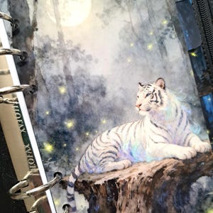 May include: A white tiger with black stripes rests on a tree stump, set against a backdrop of a moonlit forest. The tiger's fur has iridescent highlights. The image is part of a planner with silver binder rings.