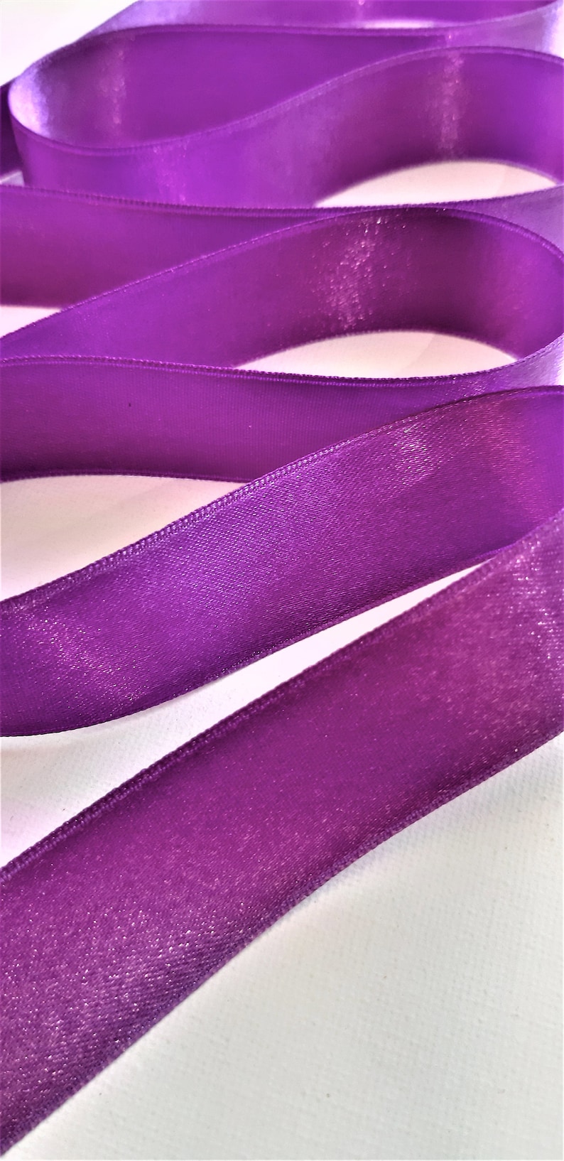 May include: Purple satin ribbon with a shimmery finish. The ribbon is folded in a wavy pattern and has a subtle sheen.