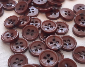 May include: A collection of brown, four-hole buttons scattered on a white surface. The buttons are round with a slightly glossy finish. Some buttons appear to be in motion, suggesting they are being poured out.