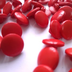 Könnte beinhalten: Eine Ansammlung leuchtend roter Knöpfe auf einer weißen Oberfläche. Die Knöpfe sind oval geformt mit einer leicht erhabenen Mitte und einer kleinen Schlaufe auf der Rückseite zum Annähen. Die Knöpfe variieren leicht in Größe und Farbton.