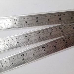 May include: Three silver metal rulers with inch markings. The rulers are stacked on top of each other. The top ruler has markings up to 25 inches, the middle ruler has markings up to 22 inches, and the bottom ruler has markings up to 20 inches.