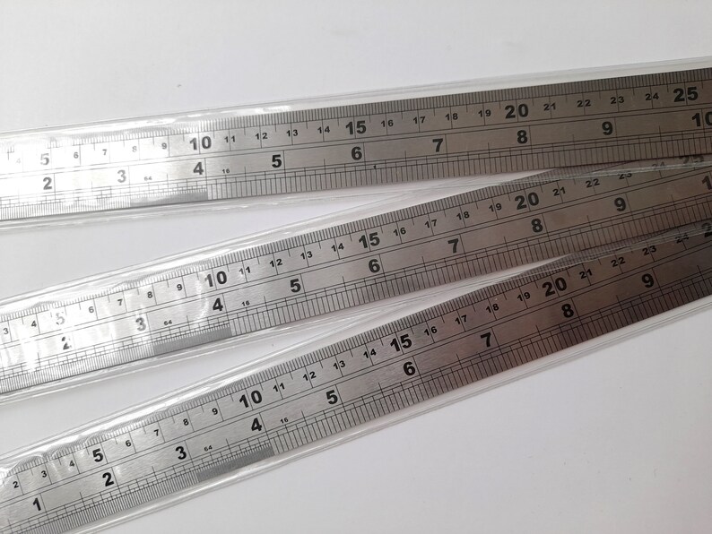 May include: Three silver metal rulers with inch markings. The rulers are stacked on top of each other. The top ruler has markings up to 25 inches, the middle ruler has markings up to 22 inches, and the bottom ruler has markings up to 20 inches.