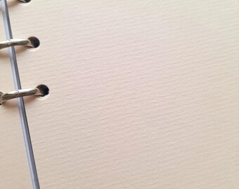 May include: Close-up of a cream-colored, textured paper notebook with silver metal ring binder. The paper has a subtle, horizontal grain. The notebook is open, revealing blank pages, perfect for writing or sketching.