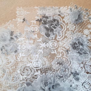May include: Close-up of delicate floral lace in grey and white. The design features intricate patterns of flowers, leaves, and geometric shapes, creating a romantic and elegant aesthetic. The lace is likely used for crafting or decorative purposes.