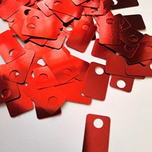 May include: A collection of small, rectangular red tags with a textured surface and a circular hole at the top. The tags are scattered across a white surface, creating a visually appealing arrangement.