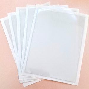 Peut inclure: Une pile de pochettes en plastique transparentes et rectangulaires, bordées de blanc. Les pochettes sont légèrement translucides, révélant le contenu. L'image est prise d'un angle légèrement surélevé, montrant la structure superposée.