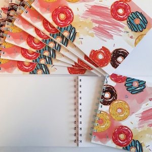 May include: A stack of spiral bound notebooks with a donut pattern. The cover features pink, red, yellow, and blue donuts on a white background with pink watercolor splatters.