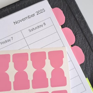 May include: A planner open to November 2025, with pink tab dividers and a sheet of pink stickers. The planner is black with a white interior. The stickers are a light pink color and have a unique shape.