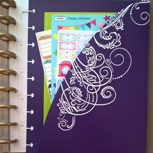 May include: A purple planner with a white decorative design on the cover. The planner has a clear pocket on the front that holds colourful stickers and a sheet of paper with the text "Happy rainbows" on it.