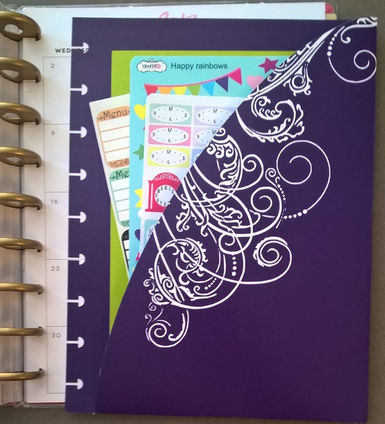 May include: A purple planner with a white decorative design on the cover. The planner has a clear pocket on the front that holds colourful stickers and a sheet of paper with the text "Happy rainbows" on it.