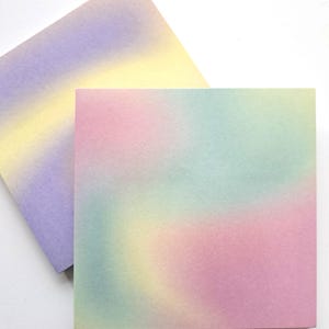 May include: Two square notepads with a soft, blended watercolor effect. One notepad features a gradient of purple, yellow, and pale yellow. The other notepad has a pastel gradient of pink, green, and yellow. The notepads are on a white background.