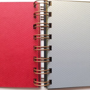 Könnte beinhalten: Ein rotes und silbernes Notizbuch mit einer Kupferdrahtbindung. Das Notizbuch hat einen strukturierten Einband.