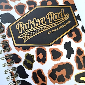 Könnte beinhalten: Ein spiralgebundenes A6 Jotta Notizbuch mit Leopardenmuster in Schwarz, Braun und Gold. Das Notizbuch hat ein schwarzes und goldenes Etikett mit der Aufschrift "Pukka Pad A6 Jotta Notebook". Ideal zum Schreiben und Notizen machen.
