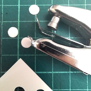 May include: A silver paper punch with a green cutting mat and three white paper circles. A white piece of paper with two holes punched in it is in the lower right corner.