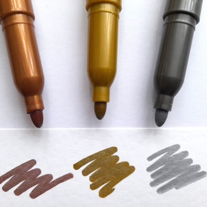 May include: Three metallic markers with brown, gold, and silver ink. The markers are on a white surface with a sample of each color drawn on a white piece of paper.