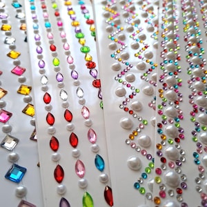 May include: Close-up of several sheets of decorative stickers. The stickers feature rows of colourful rhinestones, pearls, and various gem shapes in a variety of colours. The designs include diamond shapes, oval gems, and zigzag patterns.