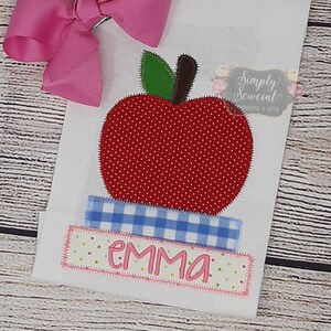 May include: A white fabric with a red apple with white polka dots, a blue and white checkered ribbon, and the name "emma" in pink letters. The fabric is decorated with a pink bow.