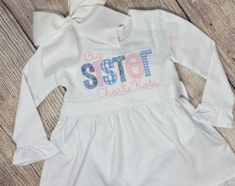 May include: White long-sleeved dress with a ruffled hem and cuffs. The dress features a pink and blue patchwork design that spells out "Big Sister" with the name "Charlie Kate" embroidered below.