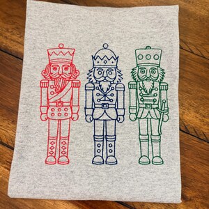 May include: A light gray folded towel featuring three embroidered nutcracker figures. The nutcrackers are in red, blue, and green, each with a unique design. The towel is made of a soft material.