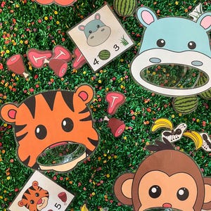 May include: A colorful educational game featuring animal-themed cutouts. The image shows a lion, tiger, hippo, and monkey, each with a circular opening. Matching cards with numbers and food items like meat and bananas are also present. The background is a green surface with small flowers.